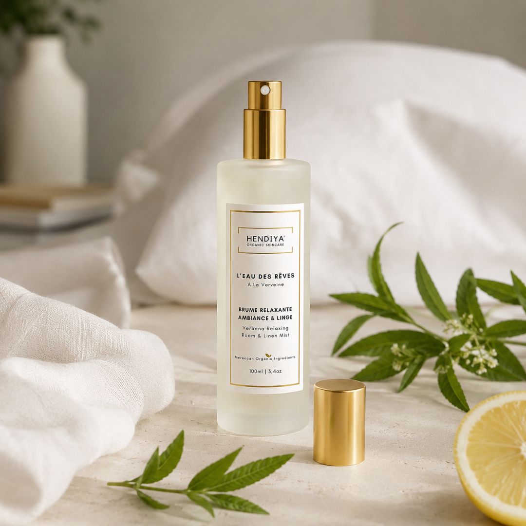 Flacon de la brume relaxante L'Eau des Rêves Hendiya entouré de brins de verveine citronnée fraîche et d'une tranche de citron, posé sur du linge de lit et des oreillers, à vaporiser en ambiance et sur le linge.