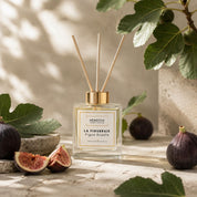 Diffuseur d'ambiance à bâtonnets de rotin La Figueraie, senteur figue royale, flacon 100ml posé avec des figues fraîches coupées et des feuilles de figuier sur une surface en pierre