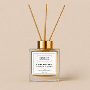 Flacon diffuseur Parfum d'Ambiance l'Orangeraie Hendiya, senteur orange épicée et écorces de cannelier, format 100ml avec ses tiges de rotin, présenté seul sur fond beige.